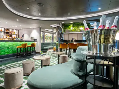 Die stilvolle Bar-Lounge an Bord der MSC Seascape zeigt eine lebendige, grün gekachelte Bar, eine gut bestückte Spirituosenwand und diverse Sitzgelegenheiten, darunter plüschige Sofas und Hocker. Ein Champagnerkühler mit Laurent-Perrier Flaschen steht im Vordergrund.