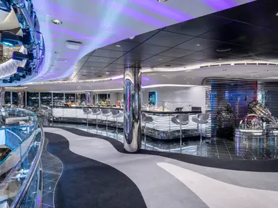 Innenansicht der Champagne & Wine Bar an Bord der MSC Seascape. Eine lange, beleuchtete Bar mit Barhockern und eine futuristische Skulptur dominieren den modernen, eleganten Raum. Violette Lichtinstallationen schaffen eine lebhafte Atmosphäre.