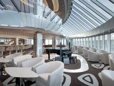 Die elegante Top Sail Lounge der MSC Seascape. Eine lange Bar, bequeme weiße Sessel, ein schwarzer Flügel und ein imposantes Glasdach bestimmen den Raum.