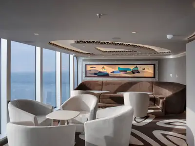 Elegante Top Sail Lounge an Bord der MSC Seascape. Bequeme Sitzgruppen, ein großes abstraktes Kunstwerk und bodentiefe Fenster mit Panoramablick auf das Meer sind zu sehen.