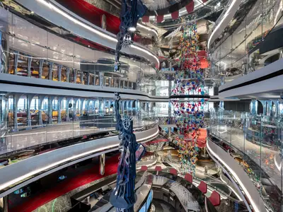 Das moderne Atrium 'Liberty Plaza' der MSC Seascape. Eine farbenfrohe Zentralskulptur, Freiheitsstatue-Replika, Glasbalustraden und eine elegante Bar sind prominent zu sehen.