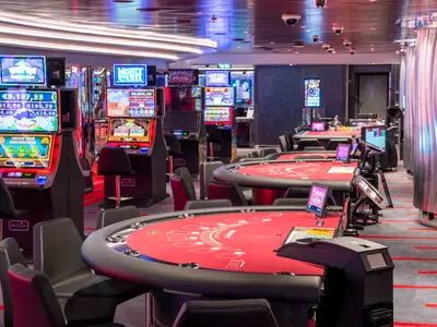 Blick in das elegant beleuchtete Casino an Bord der MSC Seascape. Zahlreiche moderne Spielautomaten und rote Spieltische stehen bereit. Schwarze Stühle sind vor den Tischen platziert.