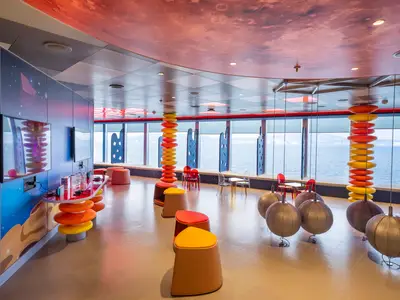 Moderner, farbenfroher Kinderbereich an Bord der MSC Seascape mit Spielekonsolen und großen Fenstern zum Meer. Orange und gelbe Stühle und Säulen schmücken den Raum.