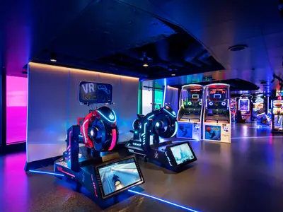 Eine futuristisch beleuchtete Hall of Games an Bord der MSC Seascape. Zwei VR Moto Rennsimulatoren mit leuchtenden Neonakzenten stehen im Vordergrund, dahinter weitere Arcade-Automaten. Ein großes Fenster mit blauer und pinker Beleuchtung ist links zu sehen. Die Szene ist ohne Personen.