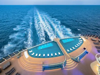 Vogelperspektive auf das offene Heckdeck der MSC Seascape mit beleuchteten Infinity-Pools, Liegen und dem weiten Meer, das die Heckwelle des Schiffes zeigt. Der Himmel ist blau, und die Beleuchtung der Pools ist prominent.