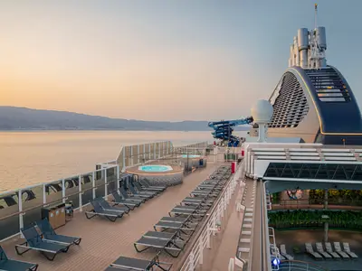 Sonnendeck der MSC Seascape im Licht des Sonnenuntergangs mit Whirlpools, Liegestühlen, Wasserrutschen, Schornstein. Ruhiges Meer, Berge im Hintergrund. Ohne Personen.