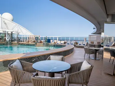Weitläufiger Poolbereich an Deck der MSC Seascape mit kühlem Wasser, bequemen Lounge-Sesseln und Tischen. Eine helle Holzterrasse führt zum blauen Meer unter strahlendem Himmel.
