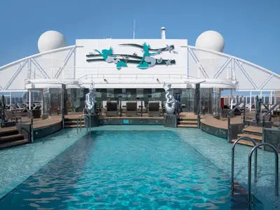 Blick auf den grossen Aussenpoolbereich des MSC Yacht Club an Bord der MSC Seascape. Ein langer Pool ist von Holzdecks, Liegen und modernen Skulpturen umgeben. Der Himmel ist klar und blau.
