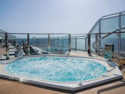 Ein blauer, sprudelnder Whirlpool auf dem Sonnendeck der MSC Seascape, umgeben von Liegestühlen und Glasgeländern mit Blick auf das glitzernde Meer unter klarem Himmel.