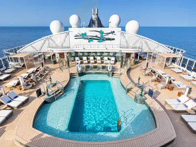 Blick auf das Pooldeck der MSC Seascape. Ein türkisblauer Pool ist umgeben von Liegestühlen auf einem Holzdeck. Im Hintergrund: weites Meer und Kunst am Schiffsaufbau.