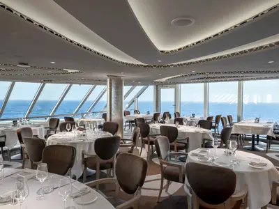 Ein elegantes Restaurant an Bord der MSC Seascape mit gedeckten Tischen, bequemen Stühlen und einem weiten Panoramablick auf das Meer durch große Fenster.