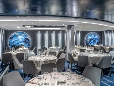 Blick in das stilvolle Aegean Restaurant an Bord der MSC Seascape. Zahlreiche Tische sind weiss gedeckt, umgeben von grauen Samtstühlen. Die Wände sind mit vertikalen, beleuchteten Paneelen und grossen, runden, blauen Kunstwerken dekoriert. Der Teppich ist blau mit weissen Mustern.