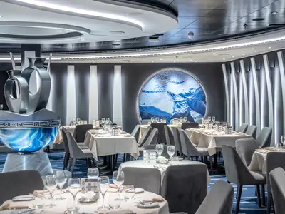 Das elegante Aegean Restaurant an Bord der MSC Seascape zeigt eingedeckte Tische und moderne, bequeme Stühle in Grau. Eine grosse, silberne Skulptur steht im Vordergrund. An der Wand prangt ein beleuchtetes rundes Bild mit einer Unterwasser-Szene und Delfinen. Der blaue Teppich ist wellenförmig gemustert.
