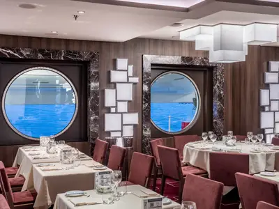 Das elegante Cala Jondal Restaurant an Bord der MSC Seascape zeigt eine Reihe gedeckter Tische und grosse Porthole-Fenster mit Blick auf das blaue Meer. Eine stilvolle Deckenbeleuchtung und Wandkunst ergänzen das moderne Ambiente.