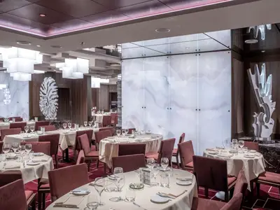 Innenansicht des eleganten Cala Jondal Restaurants an Bord der MSC Seascape. Der Raum ist mit weiss gedeckten Tischen, roten Stühlen und einem roten Teppich ausgestattet. Moderne weisse Skulpturen und hinterleuchtete Marmorwände prägen das Ambiente.