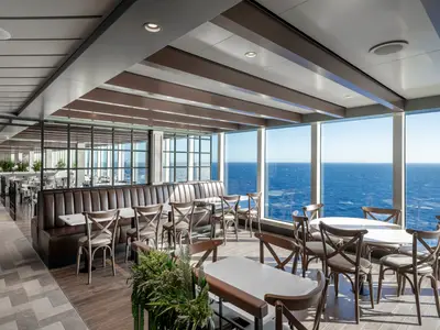 Blick in das helle Marketplace Buffet Restaurant an Bord der MSC Seascape mit Tischen, Stühlen und Sitzbänken am Fenster mit Meerblick.