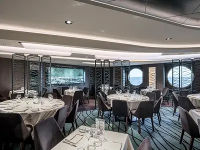 Elegantes Restaurant an Bord der MSC Seascape. Runde, gedeckte Tische, dunkle Stühle, gemusterter Teppich. Stilvolle Raumteiler und runde Bullaugen mit Meerblick.