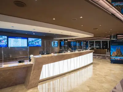 Moderne Rezeption der MSC Excursions an Bord der MSC Seascape. Beleuchteter Tresen, Bildschirme mit Ausflugsinfos, transparente Trennwände, Roll-up Banner der Sagrada Familia.