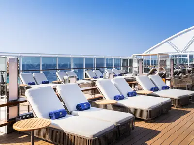 Auf dem Sonnendeck des MSC Yacht Club an Bord der MSC Seascape stehen weiße Liegen mit blauen Handtüchern. Im Hintergrund ist das blaue Meer zu sehen.