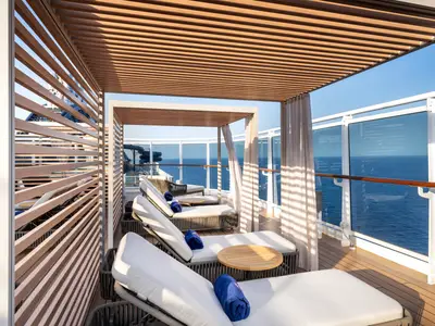 Luxuriöses Sonnendeck mit Liegestühlen, Holztischen und blauen Handtüchern an Bord der MSC Seascape. Holzlamellen spenden Schatten. Weiter Blick auf das blaue Meer und einen Wasserparkabschnitt.