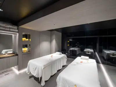 Zwei weiße Massageliegen stehen in einem modern eingerichteten Spa-Raum an Bord der MSC Seascape. Der Raum ist mit Holzverkleidungen, indirekter Beleuchtung und dunklen Spiegelflächen gestaltet. Im Hintergrund sind aufgestapelte Handtücher mit dem 'MSC AUREA SPA' Logo zu sehen.