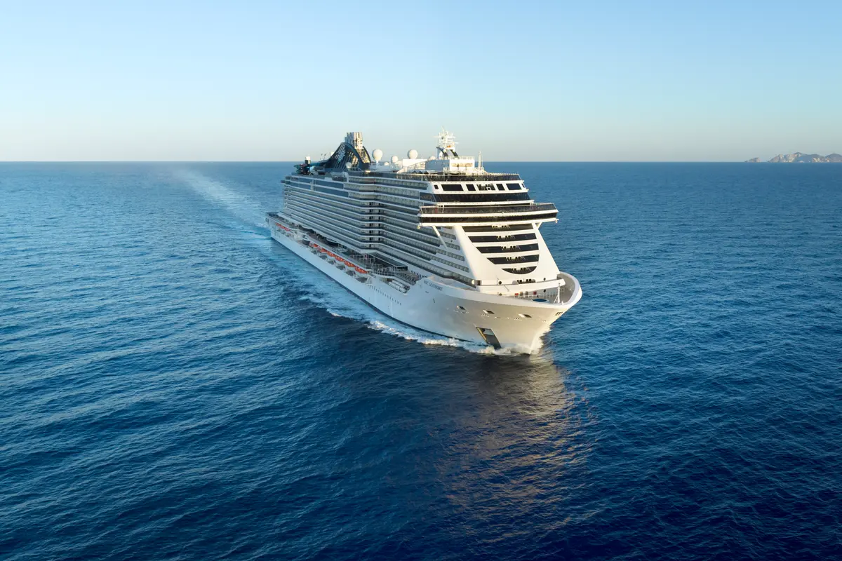 Das große Kreuzfahrtschiff MSC Seashore fährt auf blauem Meer. Luftaufnahme von schräg oben, mit Bug nach rechts. Kielspur und ferne Inseln am Horizont sichtbar.