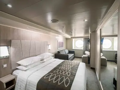 Eine helle, moderne Aussenkabine an Bord der MSC Seashore mit einem Doppelbett und weisser Bettwaesche, einem Sofa und einem runden Bullaugenfenster mit Blick nach draussen.