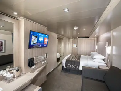 Moderne Aussenkabine an Bord der MSC Seashore. Man sieht ein Doppelbett, ein Sofa, einen grossen Flachbildfernseher und einen Schminktisch mit Spiegel und Getränken. Der Raum ist hell und einladend.