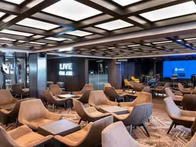 Moderne Bar-Lounge an Bord der MSC Seashore. Bequeme Sessel, Tische und eine Bühne mit Instrumenten und Leinwand. Ein Neon-Schild leuchtet 'LIVE ON AIR'.