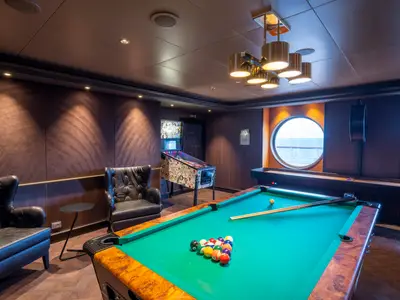 Der Poolraum an Bord der MSC Seashore zeigt einen Billardtisch, Flipperautomat und Ledersessel. Zwei Bullaugen bieten Meerblick auf den Ozean.
