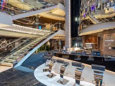 Die moderne Seashore Bar an Bord der MSC Seashore im Atrium mit einem glänzenden Tresen, Designer-Hockern und einer eleganten mehrstöckigen Treppe mit Glasgeländer und beleuchteten Stufen.