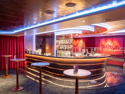 Weitwinkelansicht der modernen Bar Le Cabaret Rouge an Bord der MSC Seashore. Eine geschwungene Holztheke ist prominent im Vordergrund, dahinter beleuchtete Regale mit Spirituosen. Rote Vorhänge und eine Wanddekoration aus roten Herzen prägen den Raum. Der Boden glänzt mit integrierten Lichtlinien. Mehrere Stehtische stehen im Raum. Die Szene ist ohne Personen.