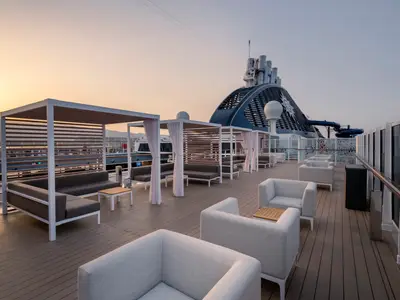 Sonnendeck der MSC Seashore im warmen Abendlicht. Moderne Lounge-Möbel und Pergolen mit Vorhängen. Im Hintergrund der Schornstein und eine Wasserrutsche.