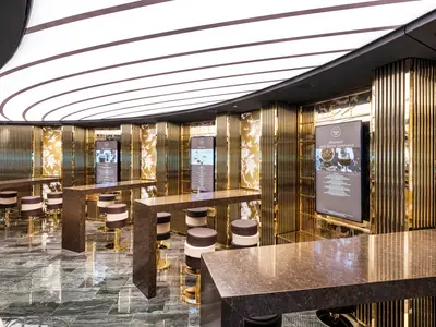 Eine luxuriöse Venchi 1878 Schokoladenbar an Bord der MSC Seashore, mit glänzenden Goldlamellen, dunklen Marmortischen und gepolsterten Hockern. Mehrere Bildschirme zeigen Angebote für Schokolade und Getränke in einem modernen Ambiente.