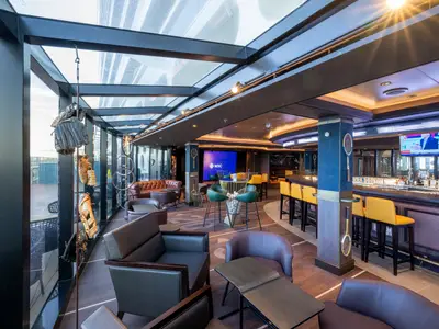 Sports Bar der MSC Seashore mit einem hellen Glasdach, einer langen Bar, gelben Hockern, Ledersesseln und Bildschirmen. Dekoriert mit Baseballhandschuhen und Tennisschlägern.
