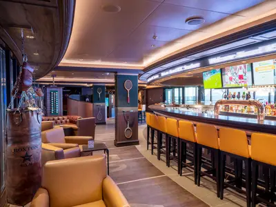 Die Sports Bar an Bord der MSC Seashore zeigt eine lange Holztheke, gelbe Barhocker und Bildschirme mit Sport. Links stehen Ledersessel und ein Boxsack, ergänzt durch Tennisschläger an den Säulen.
