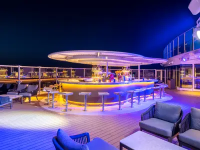 Die lebhafte Sky Bar an Bord der MSC Seashore bei Nacht, mit einer eleganten, leuchtenden Bar und Sitzgelegenheiten. Die Bar ist mit Flaschen bestückt, und die Umgebung ist in blau-violettes Licht getaucht.