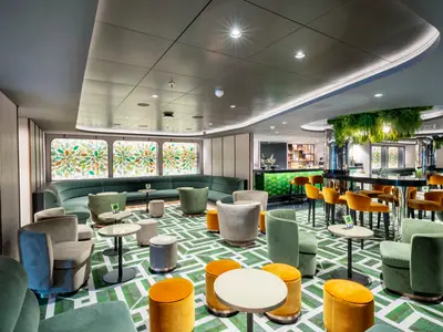 Moderne Bar-Lounge an Bord der MSC Seashore. Grüne und orangefarbene Möbel, geometrischer Boden. Grüne Bar, Deckenpflanzen und beleuchtete Fensterbilder schmücken den Raum.