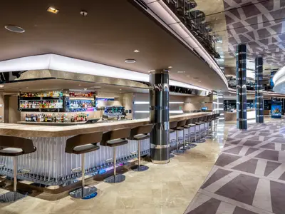 Moderne Bar an Bord der MSC Seashore. Eine lange, elegant beleuchtete Theke mit vielen Flaschen und Barhockern. Reflektierende Decken und gemusterter Teppichboden prägen das luxuriöse Interieur.