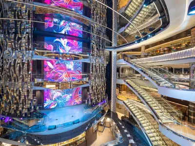 Das mehrstöckige Atrium der MSC Seashore besticht durch große LED-Bildschirme mit floralen Motiven, glänzende Spiegelwände und elegante Treppen mit Glasgeländern. Eine moderne Bar ist ebenfalls sichtbar.