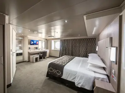 Geräumige, barrierefreie Aussenkabine an Bord der MSC Seashore. Das Bild zeigt ein gemachtes Doppelbett, einen Schreibtischbereich mit Stuhl und TV, sowie runde Bullaugenfenster und einen Ganzkörperspiegel.