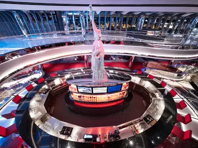 Vogelperspektive auf die moderne Liberty Plaza mit einer grossen Statue der Freiheitsstatue und einer zentralen Bar auf der MSC Seashore. Das Atrium zeigt mehrere Ebenen mit reflektierenden Oberflächen und roten Sitzen.