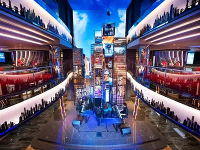 Zwei Pianisten spielen an Flügeln im mehrstöckigen, hell beleuchteten Times Square Atrium der MSC Seashore, umgeben von digitalen Skylines, Geschäften und Bars. Eine lebhafte Szene.