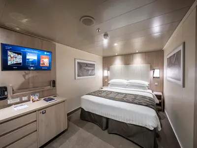 Helle Innenkabine an Bord der MSC Seashore. Ein großes Doppelbett mit zwei Kissen und einer gemusterten Decke dominiert den Raum. Links ein Schreibtisch mit TV und Telefon, rechts ein gerahmtes Bild über dem Bett.