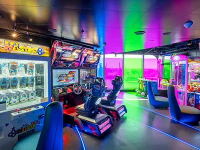Belebte Spielhalle, auch Hall of Games genannt, an Bord der MSC Seashore. Zahlreiche Arcade-Automaten, darunter Rennspiele und Key Master, erstrahlen in neonfarbenem Licht. Der Raum wird von pinken, blauen und grünen Lichtreflexen durchflutet.