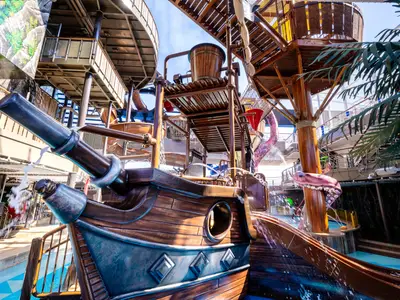 Der Pirates Cove Wasserpark an Bord der MSC Seashore. Eine piratenschiffartige Struktur mit Wasserrutschen, Kanonen und einem großen Kippeimer. Grünzeug und Schiffsdecks im Hintergrund.