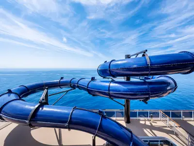 Detailansicht der leuchtend blauen Wasserrutsche des Pirates Cove Aquaparks an Bord der MSC Seashore. Die Rutsche schlängelt sich über dem teakfarbenen Deck, mit weitem Blick auf das blaue Meer und den bewölkten Himmel im Hintergrund.