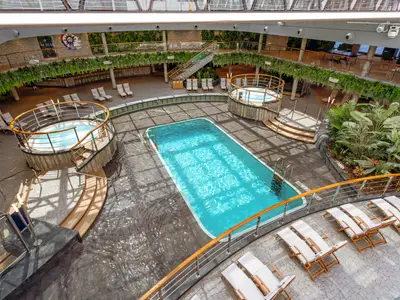 Vogelperspektive auf den Jungle Pool an Bord der MSC Seashore. Ein grosser Pool, zwei Whirlpools, zahlreiche Liegen und eine tropische Bepflanzung auf mehreren Decks sind zu sehen.