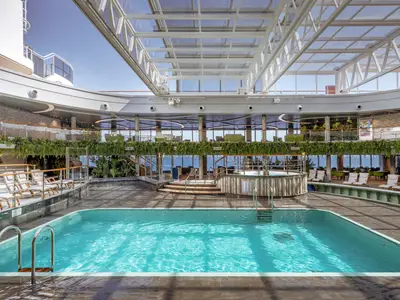 Blick auf den grossen, hellblauen Pool der Jungle Pool Lounge an Bord der MSC Seashore. Rundherum Liegen, ein Whirlpool und grüne Pflanzen. Ein Glasdach sorgt für Tageslicht.