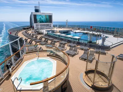 Sonniges Deck der MSC Seashore mit großem Pool, Liegestühlen, Whirlpool und Bildschirm. Im Hintergrund ist das weite blaue Meer mit der Schiffspur sichtbar.